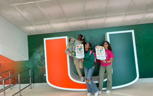 Three older Girl Scouts holding up a vest plus UM posters in the University of Miami U at Cookie U